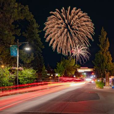 Una larga exposición de luces de cola rojas en The Village en Big Bear Lake. Fuegos artificiales del 4 de julio son visibles en el cielo nocturno.