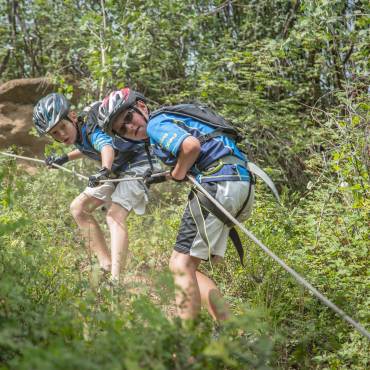 Dos niños, equipados con material de seguridad, bajan por la ladera de una colina con ayuda de una cuerda de escalada; a su alrededor se extiende un frondoso bosque.