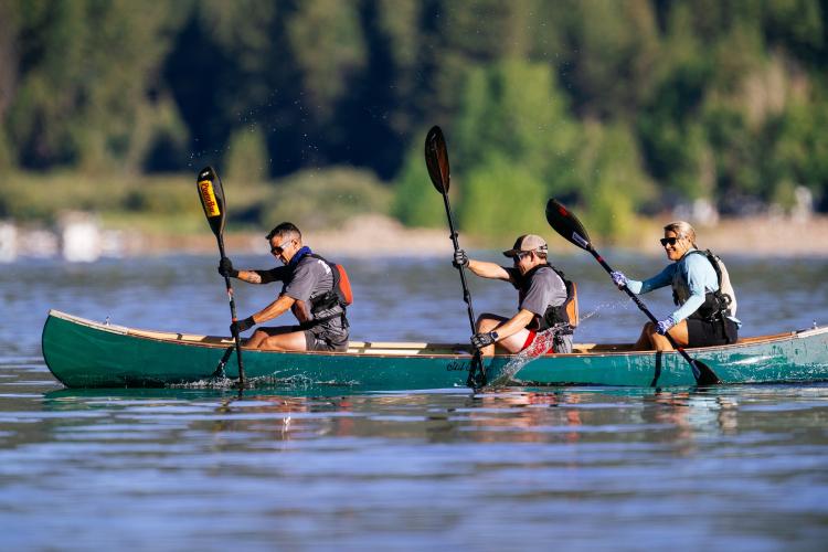 A group of three on a kayak during LakeFest, working together to row with their paddles, splashing water upwards as they go.