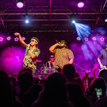 Una foto tomada entre el público del ciclo de conciertos de verano, los cuatro miembros de la banda tocando sus instrumentos y cantando.