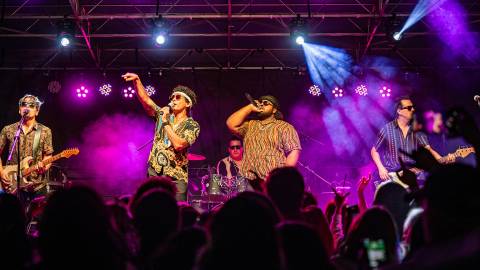 Una foto tomada entre el público del ciclo de conciertos de verano, los cuatro miembros de la banda tocando sus instrumentos y cantando.