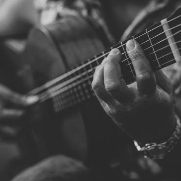 Foto en blanco y negro de alguien que sostiene y toca una guitarra acústica; la mano más alejada sostiene una púa de guitarra.