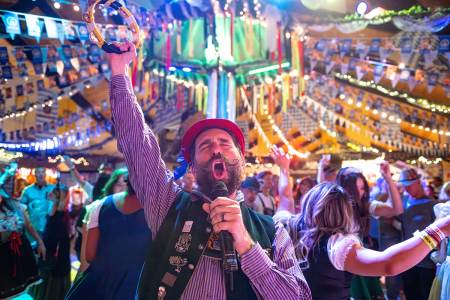Un cantante vestido con el traje tradicional alemán del Oktoberfest aparece cantando ante un micrófono; detrás de él hay una gran multitud.