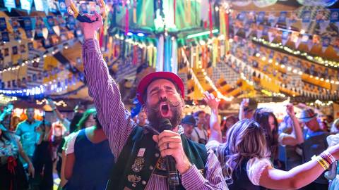 Un cantante vestido con el traje tradicional alemán del Oktoberfest aparece cantando ante un micrófono; detrás de él hay una gran multitud.