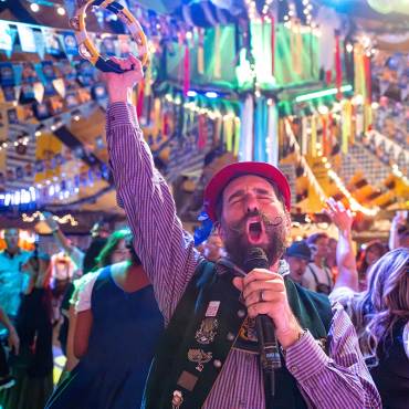 Un cantante vestido con el traje tradicional alemán del Oktoberfest aparece cantando ante un micrófono; detrás de él hay una gran multitud.