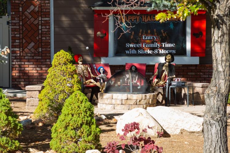 Una exposición de espantapájaros y Día de los Muertos de 2025: hay esqueletos descansando en sillas rojas junto a una hoguera.