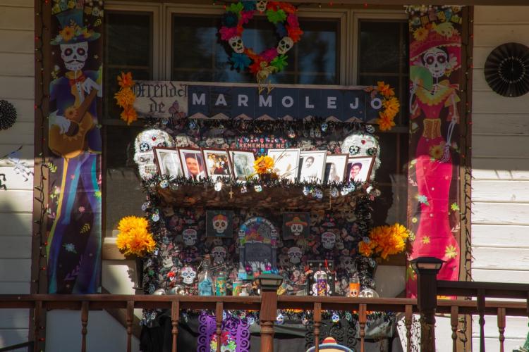A 2025 Scarecrow & Dia de Los Muertos Display - la ofrenda tiene fotos de personas, velas, esqueletos y flores.