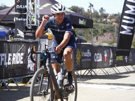 A photo of female cyclist Siena Hermon cheering with one hand raised in the air after crossing the finish line at a bike race