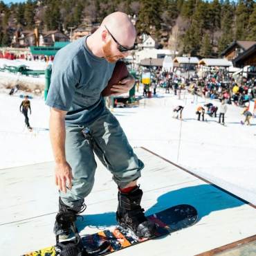 Foto de un esquiador de snowboard sosteniendo un balón de fútbol americano; al fondo se ve una multitud en la base de la estación de esquí Bear Mountain.