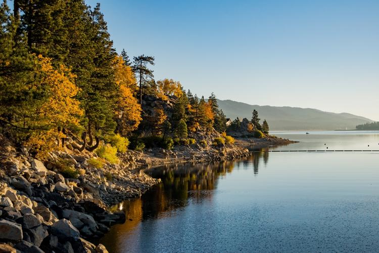 Una foto tomada a orillas del lago Big Bear, con la puesta de sol iluminando suavemente los árboles teñidos de colores otoñales y la orilla de la playa.