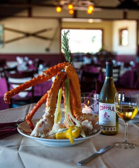 A photo of a crab leg dish at the Pines Lakefront Tavern, beside it is a bottle of Coppola Chardonnay and a filled wine glass