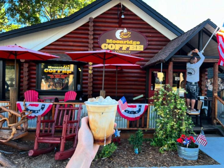 A photo of a hand holding out an iced coffee in front of the Moonridge Coffee Co building.