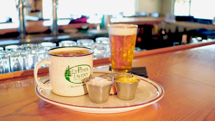 Una foto de cerca del Pines Tavern Cup O' Tri-Tip Chili, hay cebollas, queso cheddar, Tri-Tip, y una taza de chili.