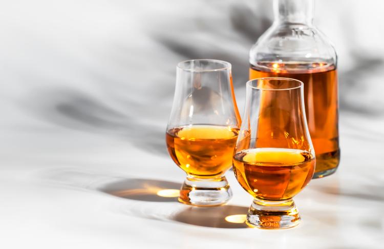 Un decantador y dos vasos están llenos de bourbon de color ámbar. Proyectan sombras y reflejos sobre el fondo blanco.
