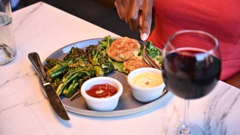 La mano de una mujer corta comida en un plato de un restaurante de Big Bear Lake. Junto al plato, un vaso de vino tinto.
