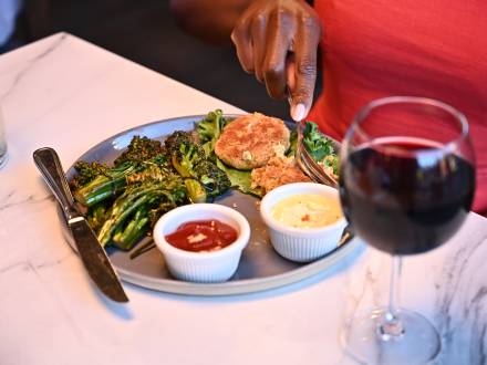 La mano de una mujer corta comida en un plato de un restaurante de Big Bear Lake. Junto al plato, un vaso de vino tinto.