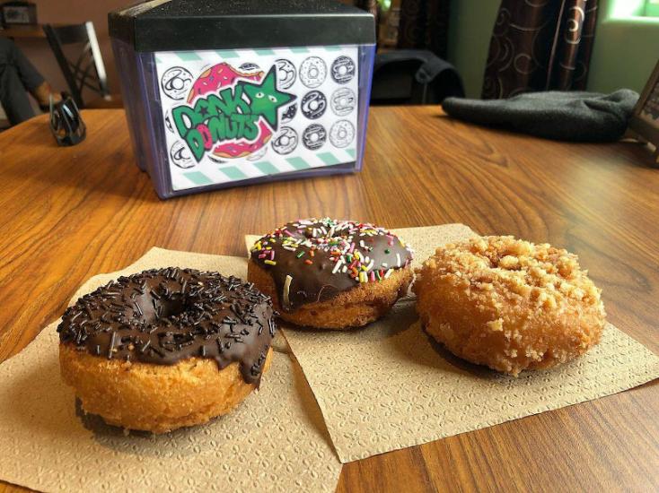 Tres donuts sobre una servilleta en una mesa. En el fondo, un servilletero con el logotipo de Dank Donuts.