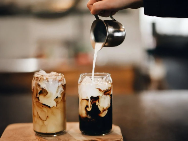 Se está vertiendo leche al vapor en dos vasos de la altura aproximada de una lata de refresco; el café fuerte que hay dentro se está mezclando con la leche.