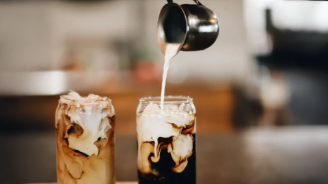 Se está vertiendo leche al vapor en dos vasos de la altura aproximada de una lata de refresco; el café fuerte que hay dentro se está mezclando con la leche.