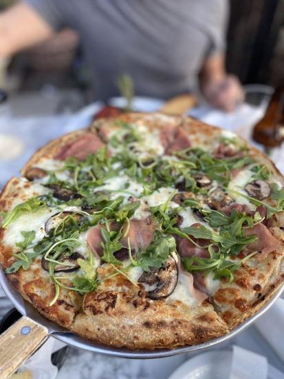 A top-down photo of a pizza from La Pergola Trattoria, it has arugula, mushrooms, and sliced prosciutto.