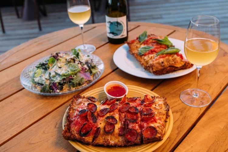 Una foto de la pizza y el vino de De La Nonna: se ve una ensalada en un plato transparente y pizzas cuadradas con diferentes ingredientes.