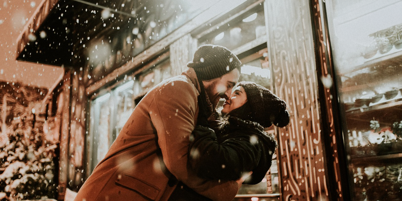 Una foto de una pareja abrazándose con cariño, sonriendo delante de una tienda mientras la nieve cae a su alrededor.