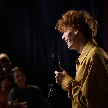 Foto de un hombre con una camisa amarilla sosteniendo un micrófono, sonriendo alegremente mientras habla frente a una multitud.