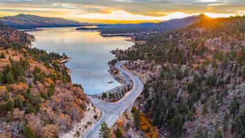 Una toma con dron del lago Big Bear en otoño, con la puesta de sol asomando por detrás de las montañas y reflejándose suavemente en el lago.