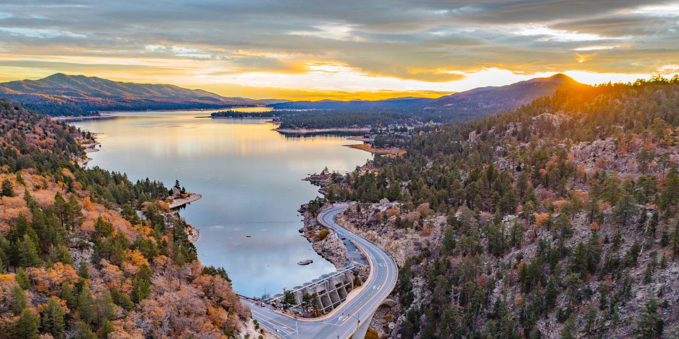 Una toma con dron del lago Big Bear en otoño, con la puesta de sol asomando por detrás de las montañas y reflejándose suavemente en el lago.