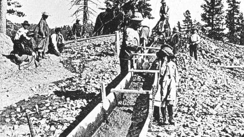 A black and white photo of prospectors panning for gold using a man made stream.