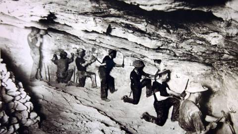 Fotografía en blanco y negro de un grupo de buscadores de oro en una cueva, utilizando cinceles para buscar en la roca.