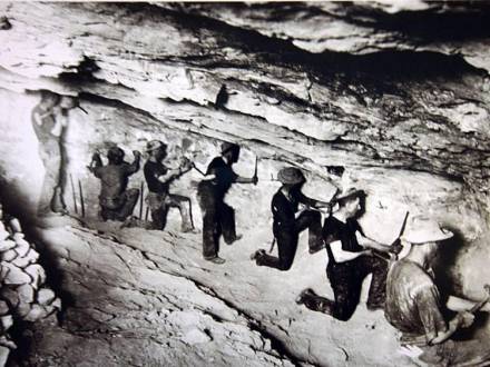 Fotografía en blanco y negro de un grupo de buscadores de oro en una cueva, utilizando cinceles para buscar en la roca.