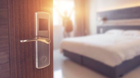 Un primer plano de la entrada de una habitación de hotel, desenfocado al fondo hay una cama y una persona de pie abriendo las cortinas.