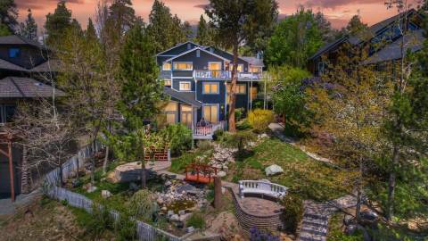 Una casa azul marino de tres plantas situada en lo alto de una colina cubierta de exuberante vegetación; detrás de ella se divisan el cielo teñido por la puesta de sol y el bosque.