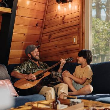 Un padre y un hijo sentados en el sofá de su casa; el padre sostiene una guitarra acústica grande y sonríe.