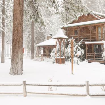 La nieve cubre el patio de una cabaña en Big Bear, con altos árboles que rodean la alta cabaña.
