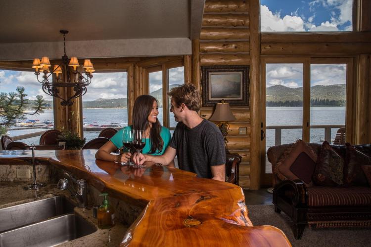 Una pareja se sienta en un mostrador de madera con copas de vino en el interior de una cabaña con vistas al lago Big Bear por las ventanas.