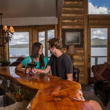 Una pareja se sienta en un mostrador de madera con copas de vino en el interior de una cabaña con vistas al lago Big Bear por las ventanas.