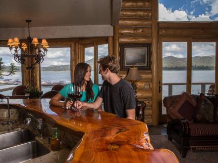 Una pareja se sienta en un mostrador de madera con copas de vino en el interior de una cabaña con vistas al lago Big Bear por las ventanas.